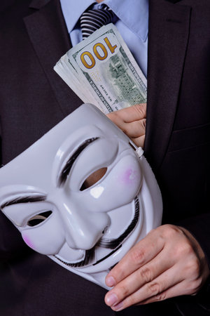 A mysterious person in formal wear holds an anonymous mask and pile of us dollars. Giving a bribe, corruption, giving money to politicians conceptsの写真素材