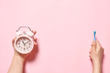 Morning concept, pink alarm clock and a toothbrush in the hands of a young woman on a pink background. Flat layの写真素材