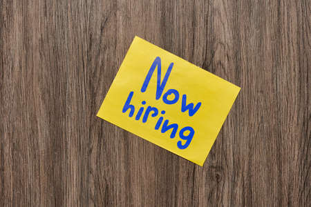 Now hiring text on yellow note with blue sheet on wooden desk. Conseptの写真素材