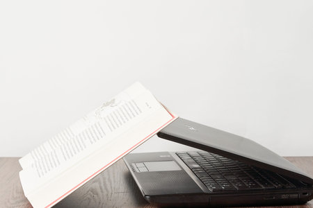 an open book and a laptop on a wooden table with a white wall in the background behind it is a black...