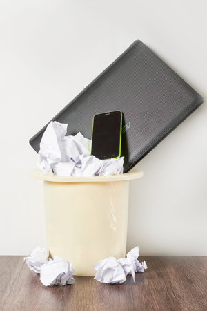 a cell phone in a trash can with crumpled paper on the floor next to it is an open laptop computer