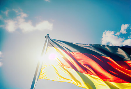 a german flag flying high in the blue sky with white clouds and bright sun shining through its rays. Generative AIの素材