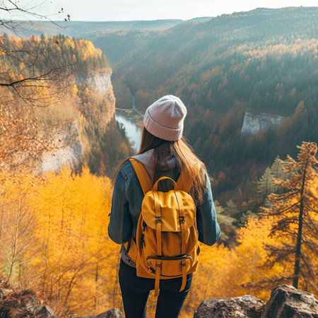 a woman wearing a yellow backpack standing on a cliff overlooking the mountains and trees in autumn, looking out to the valley belowの素材