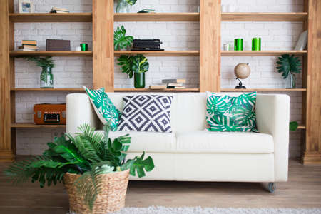 Living room Comfortable interior. Decorative pillows on a white sofa, bookshelves, decor with tropical leaves. Horizontal photo of home furniture.の写真素材