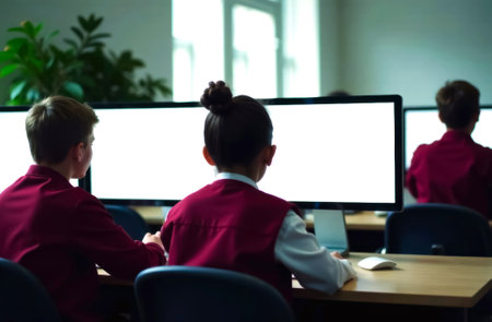 schoolchildren boy and girl in red uniform sitting at desk in classroom in front of computer monitor, computer monitor mockup, close-up from backの素材