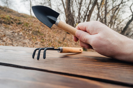 a hand holds a small garden trowel, and a miniature rake is lying next to it, both tools with wooden...