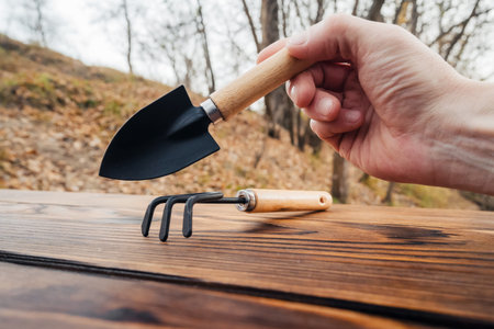 close-up of a hand holding a small garden trowel raised above a small rake, which is lying on a brown wooden table in an autumn park, emphasizing the process of planting and careの写真素材