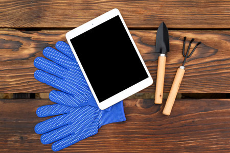 white tablet lies on blue work gloves, and next to them are a trowel and a weeder with wooden...