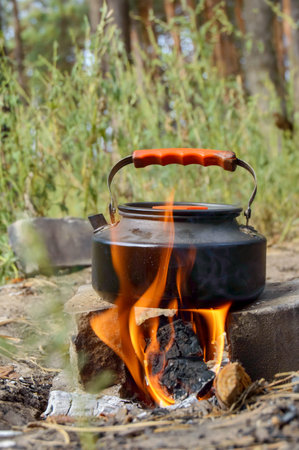 a vertical frame featuring a black camping kettle with an orange handle, set on bricks over a...