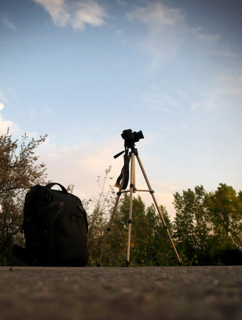 black backpack and a camera on a tripod on the ground or stone, their silhouettes against the bright evening sky with clouds and dark tree crowns, preparation for a photo shootの写真素材