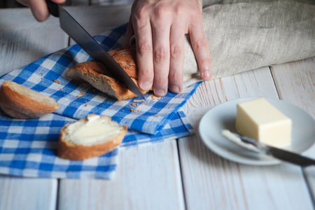 Traditional breakfast cooking. Hands of man cut a baguette over the table with bread and napkin....