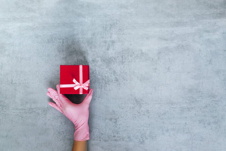 Love gift. Valentine day greeting. protection. Hand in medicine glove holding red present box on gray copy space. Advertising background.の写真素材