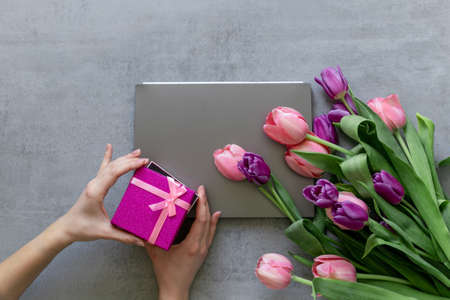 A woman's hands open a pink gift box. A gray laptop and pink tulips on a gray concrete background.の写真素材