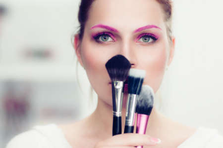 The makeup artist with bright pink make-up holds powder brushes. She covers with them the face. Studio portrait.の写真素材