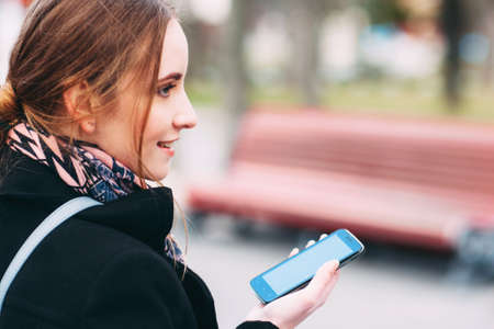 Young attractive woman in a black coat is walking in a park with blank mobile phoneの写真素材