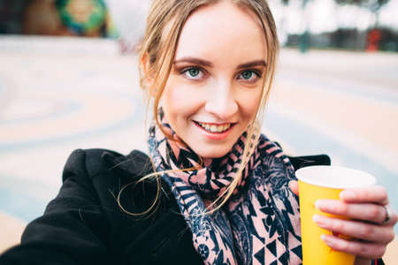 Young attractive woman in a black coat with a cup of coffee taking selfie in a parkの写真素材