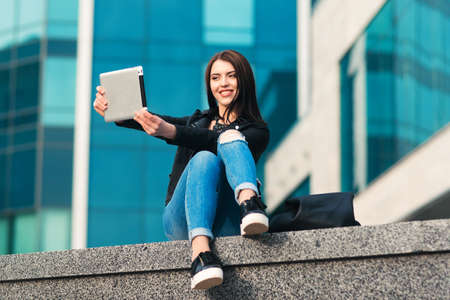 Smiling girl in a black jacket and blue jeans takes selfie with a silver tablet on the city backgroundの写真素材