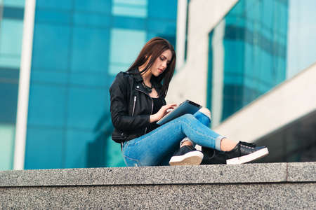 Young attractive woman in a black jacket and blue jeans surfing on a silver tablet against urban city backgroundの写真素材
