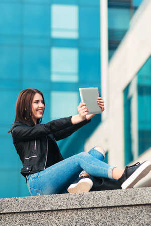 Smiling girl in a black jacket and blue jeans takes selfie with a silver tablet on the city backgroundの写真素材