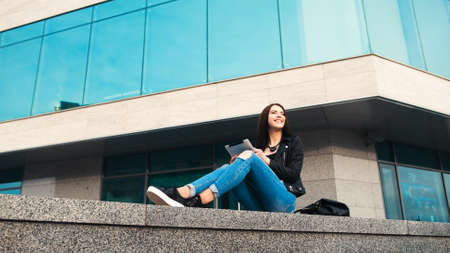 Smiling girl in a black jacket and blue jeans sitting with a silver tablet on the city backgroundの写真素材