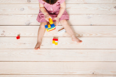 little girl in red dress sitting on the wooden floor and playing Lego. Top view