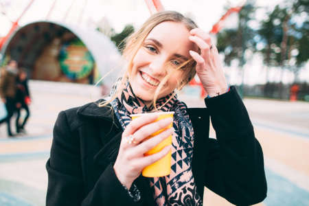 Smiling young attractive woman in a black coat is walking in a park with a cup of coffee. she openly laughs and enjoysの写真素材