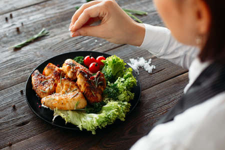 Preparation of chicken wings on wooden table. Chief cooks grilled spicy chicken with fresh vegetables. Over the shoulder view of food preparation.の写真素材