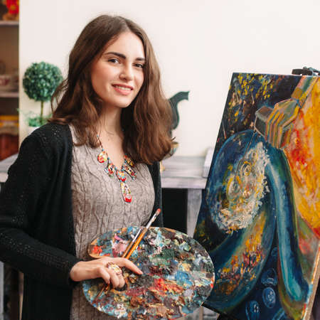 Creative happy painter paints a colorful picture. Closeup of painting process in art workplace. Smiling positive woman painter paints in her studio.の写真素材