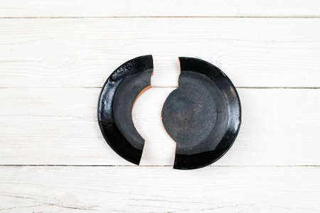 Shards of a broken black plate on white wooden surface.  Problem, accident, divorce, misfortune, scandal, quarrel concept. Flat lay of contrast black broken on two parts plate.の写真素材