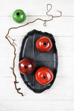 Many ceramic red pomegranates and black plate on white wooden background. Flat lay set of color  house decor. Organic food, pottery, decoration concept. Bright ceramic red pomegranates on black dishの写真素材