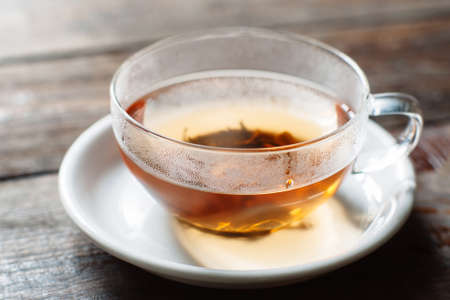Cup of hot black tea close-up. Glass cup on saucer front view. Cup with tea leaves on dark wooden background.の写真素材