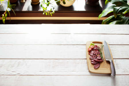 Sliced salami and tomato on cutting board with knife on white wooden table. Ready for serving cutting board with sliced sausage. Free space. Blured backgroundの写真素材