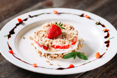 Layered strawberry cake with cream on white plate closeup. Front view on layered cake with cream, strawberry and cookies on white plate, decorated with chocolate sauce and leaves of fresh mint.の写真素材
