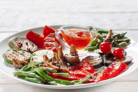 Grilled vegetables with red sauce on white plate. Front view on grilled asparagus, vegetable marrow, tomato, pepper, eggplant and mushrooms on white plate with cup of ketchup close-upの写真素材