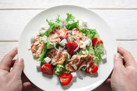 Serving plate with steaks, strawberry and feta cheese flat lay. Top view on hands, holding plate with original salad with chicken steaks, strawberries, onion and feta cheese on white wooden backgroundの写真素材