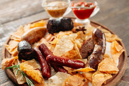 Beer snacks set on wooden plate. Closeup of wooden catering platter with assorted snacks for beer with two sauces. Different sausages and chips on wooden background.の写真素材