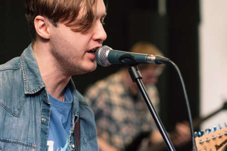 Guy singing into the microphone close-up. Profile of emotionally singing man on stage, guitar player on blurred background. Vocalist in jeans coat with closed eyes sings.の写真素材