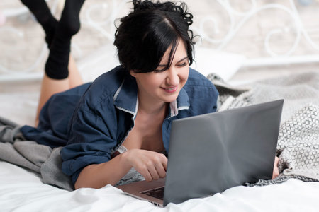 Smiling woman on bed with laptop. Front view on smiling woman lying down the bed in front of her...