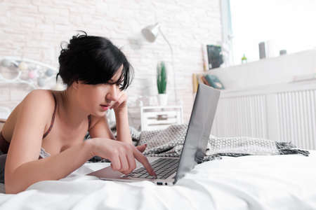 Nude brunette working on laptop close-up. Head and shoulders of pretty brunette woman, lying on bed in undies and working on laptop. Lying on bed brunette in lingerie with computerの写真素材