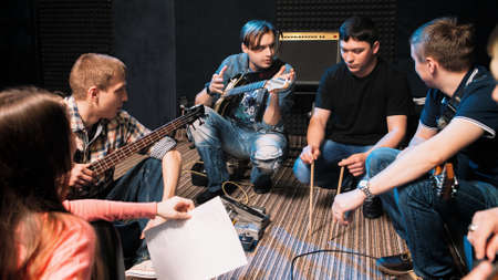 Music band discussing drummers party in song. Front view on young musicians sitting on the floor, everyone looking at guy with drumsticks.の写真素材