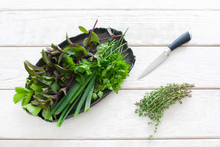 Plate with flavoring, knife and thyme on white flat lay. Top view on black clay plate with basil, parsley and chives, bunch of thyme and knife on white wooden backgroundの写真素材