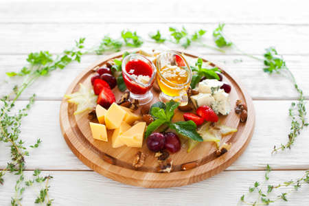 Plate with snacks for wine in herbal frame on white. Top view on few kinds of cheese and fruits on catering platter with red and yellow sauces. Wine snacks plate on white wooden backgroundの写真素材