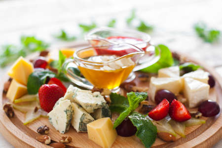 Wine snacks set on wooden cutting board closeup. Fruits and different kinds of cheese with yellow and red sauces on round wooden board. Strawberry, grape, nuts and cheeseの写真素材