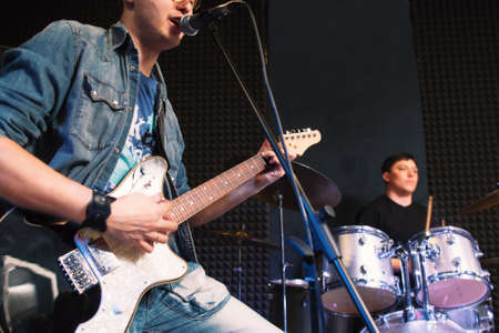 Playing on guitar vocalist and drummer on background. Focus on guitar player singing into the micriphone, playing drummer on background. Student music band on stageの写真素材