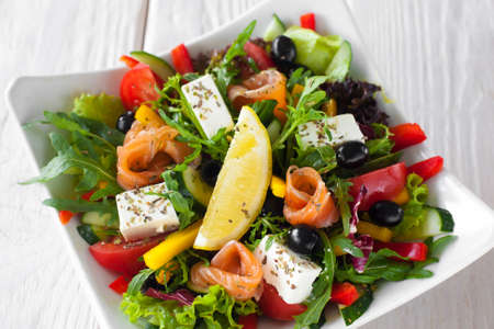 Mediterranean salad with salmon and brie top view. Appetizing salad with salmon, brie, olives and vegetables, served with lemon slice in white bowl on wooden table.の写真素材