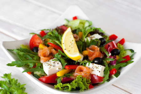 Salmon salad traditional serving with lemon on white wooden background. Top view on white bowl with smoked salmon salad, pepper, brie, olives, lettuce, decorated with slice of lemon.の写真素材