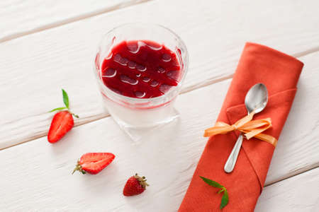 Strawberry organic panna cotta in glass with spoon flat lay. Dessert panna cotta with fresh strawberry slices served in glass and spoon on orange napkin on white wooden tableの写真素材