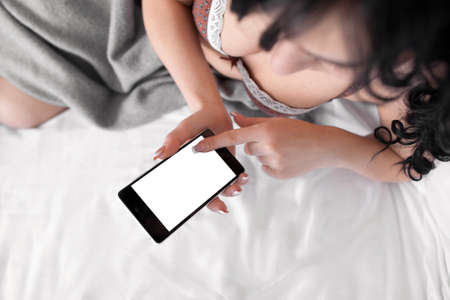 Naked brunette touching cell phone with empty screen. Top view on nude woman in bed using her smartphone. Mobile phone screen with copyspaceの写真素材