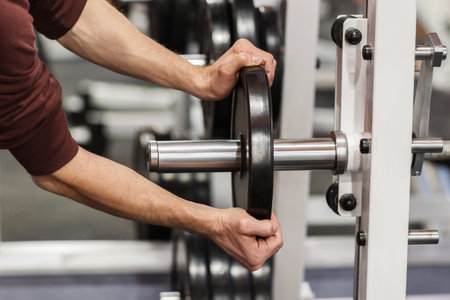 Male hand putting weight plate on barbell. Weightlifting training preparation. Weight plate in...