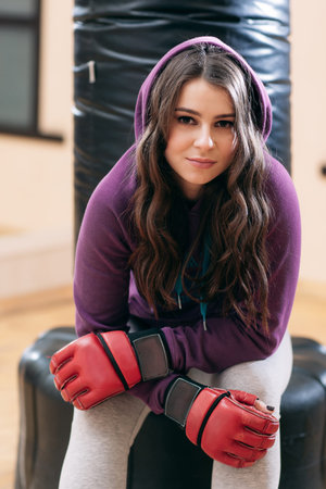 Young girl in sport wear, sitting on punching bag in gym . Pretty female athlete in red boxing...